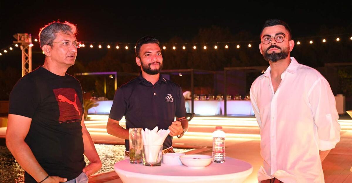 Sukesh P M, centre, with Sanjay Bangar on the left and Virat Kohli on the right at an outdoor venue in the evening.